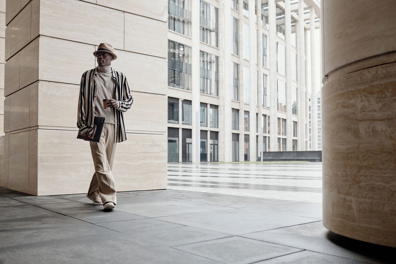 about-01 Fashionable man wearing hat and striped jacket walking in a modern cityscape.