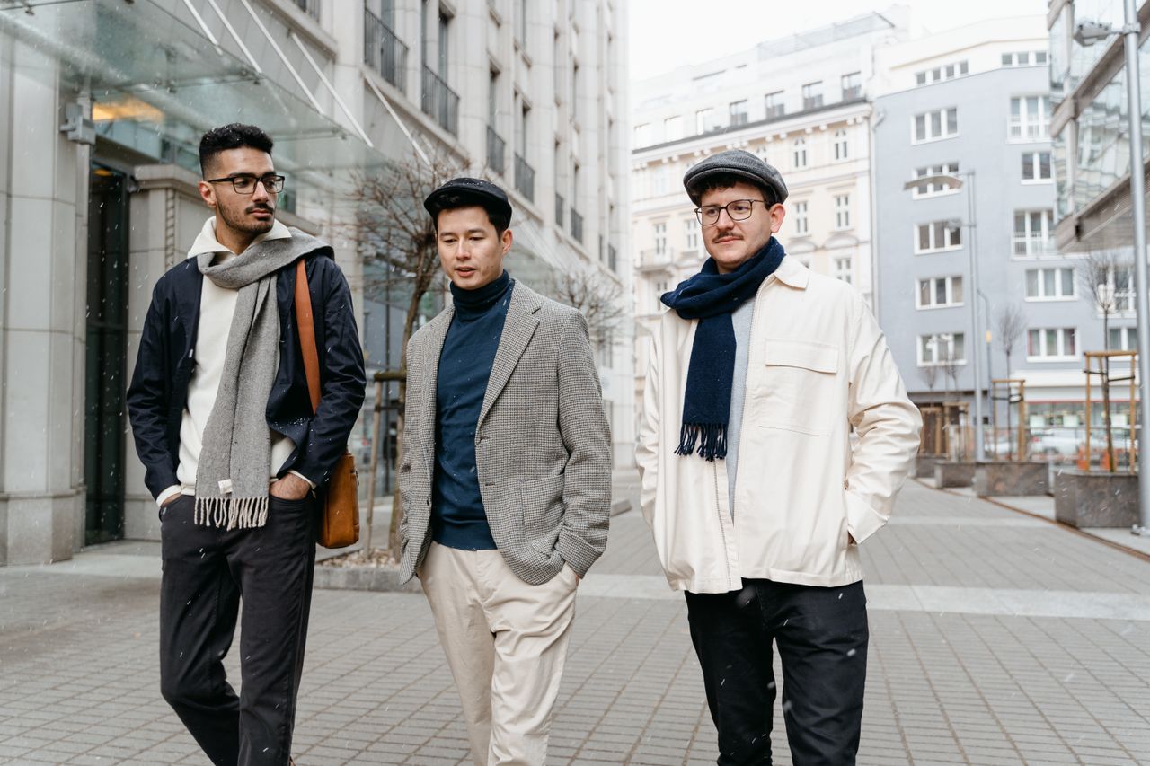 hero-img-01 Three fashionable men stroll along a city street, showcasing modern winter style with scarves and flat caps.
